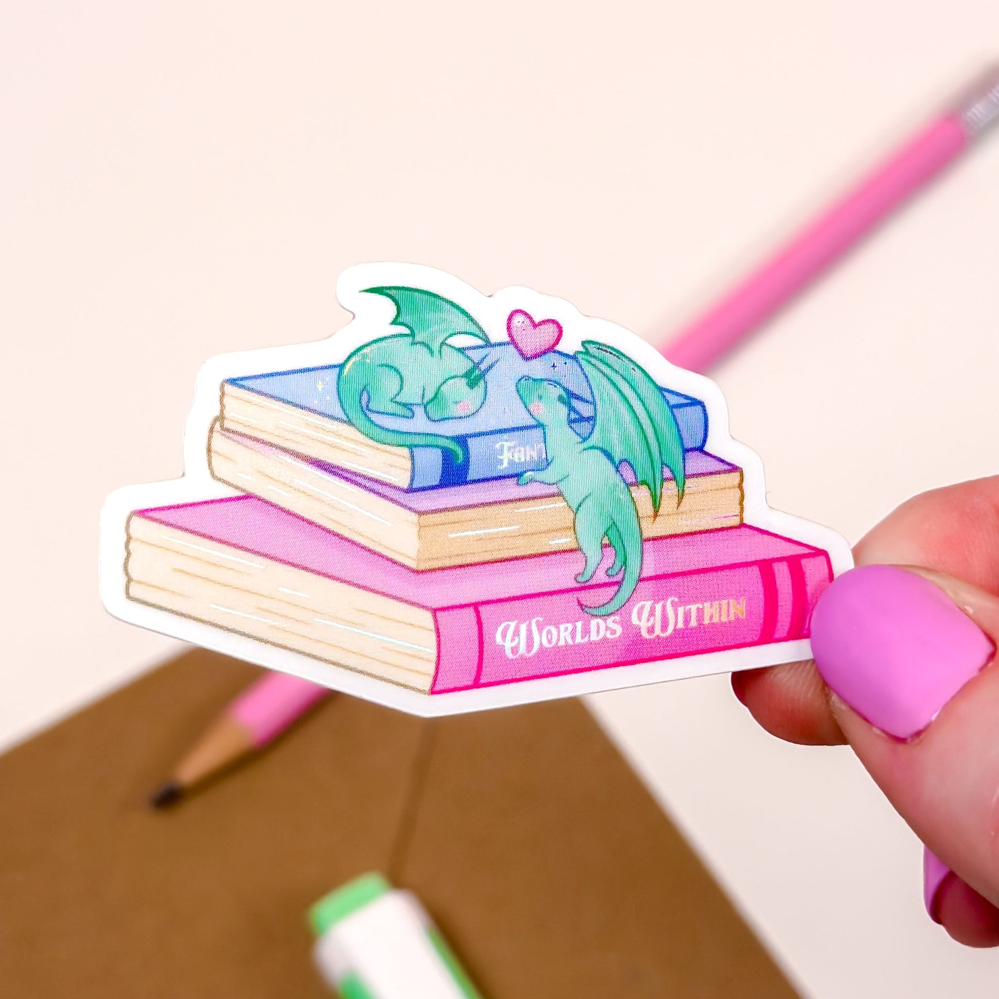 Sticker of dragons on books held by a hand with pink nail polish, with a pencil and marker in the background.