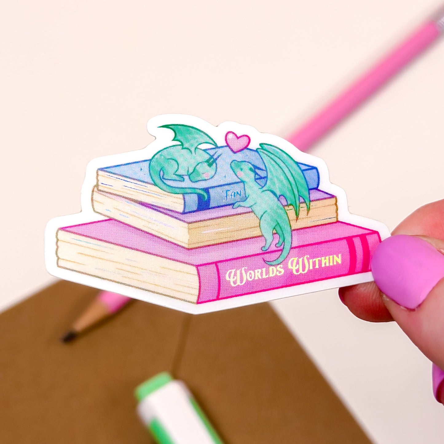 Sticker of books with dragons on a pink background held by a hand with pink nail polish.